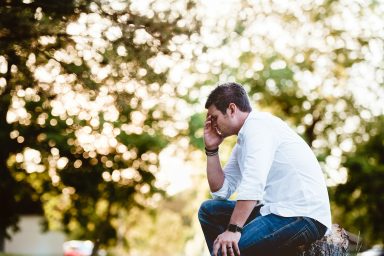 Hombre pensativo sentado en un tronco, rodeado de árboles y luz suave del atardecer.